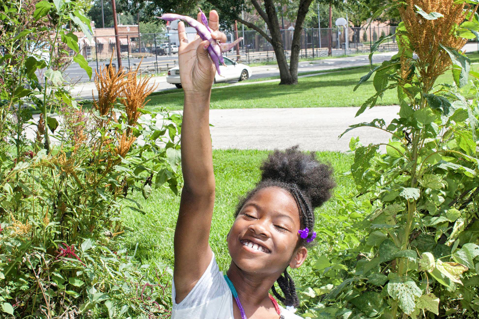 A young girl standing in a garden smiles wide holding up two purple beans