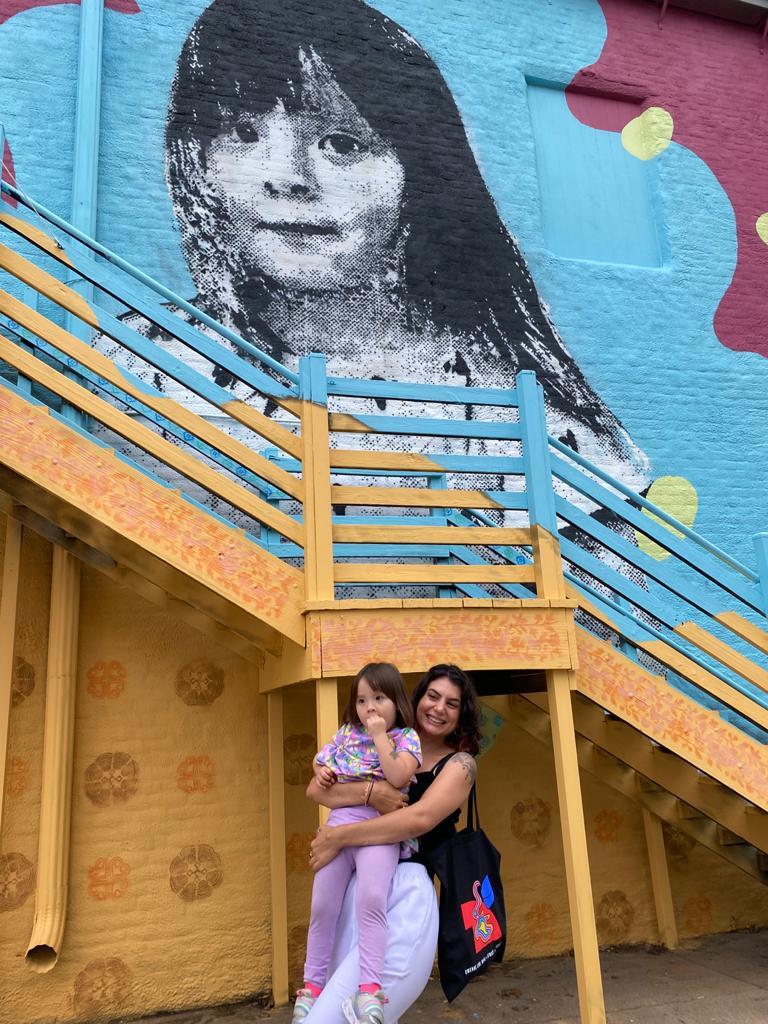 Artist Oria Simonini Hold A Child In Front Of A Mural Of Their Likeness