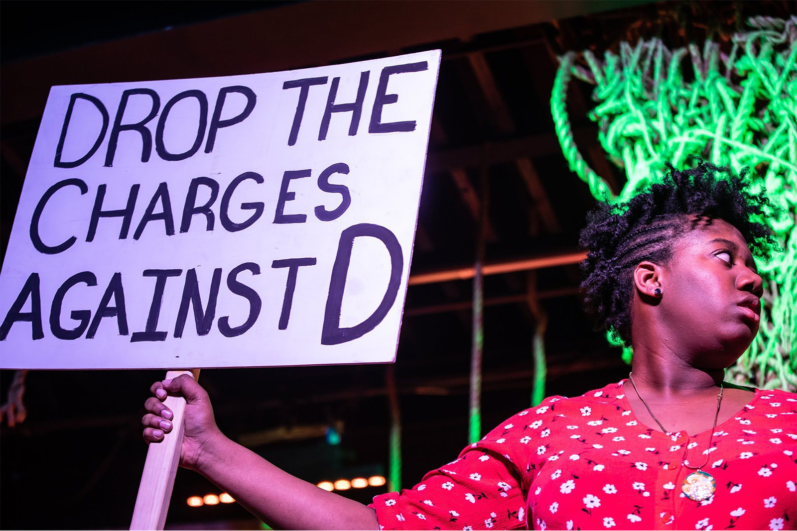 Blood at the Root Actress looks out of frame wearing a worried look and holding a protest sign that reads Drop the Charges Against D