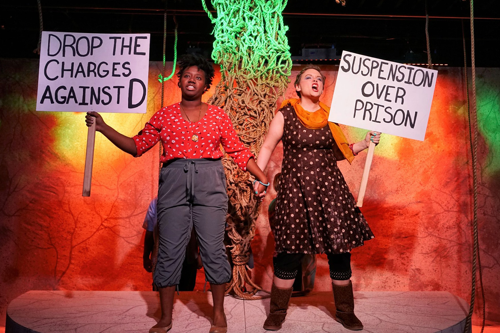 Blood at the Root Two actresses stand center stage holding hands shouting in protest Both hold a protest sign reaading Suspension Over Prison and Drop the Charges Against D