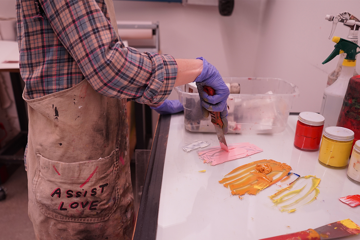 Co-Op-Program - An artist mixes printmaking ink with a knife, the artist wears a paint splattered apron with the words "Assist Love" written on the pocket.