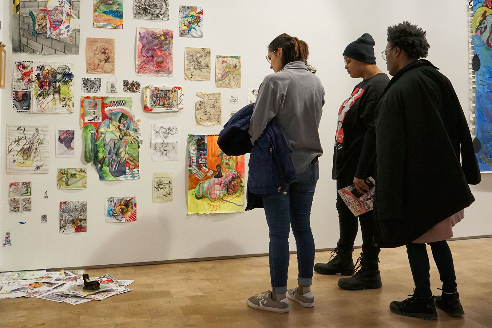 Exhibition Three people stand in the gallery looking at several small works by Thalia Rodgers assembled together on that wall