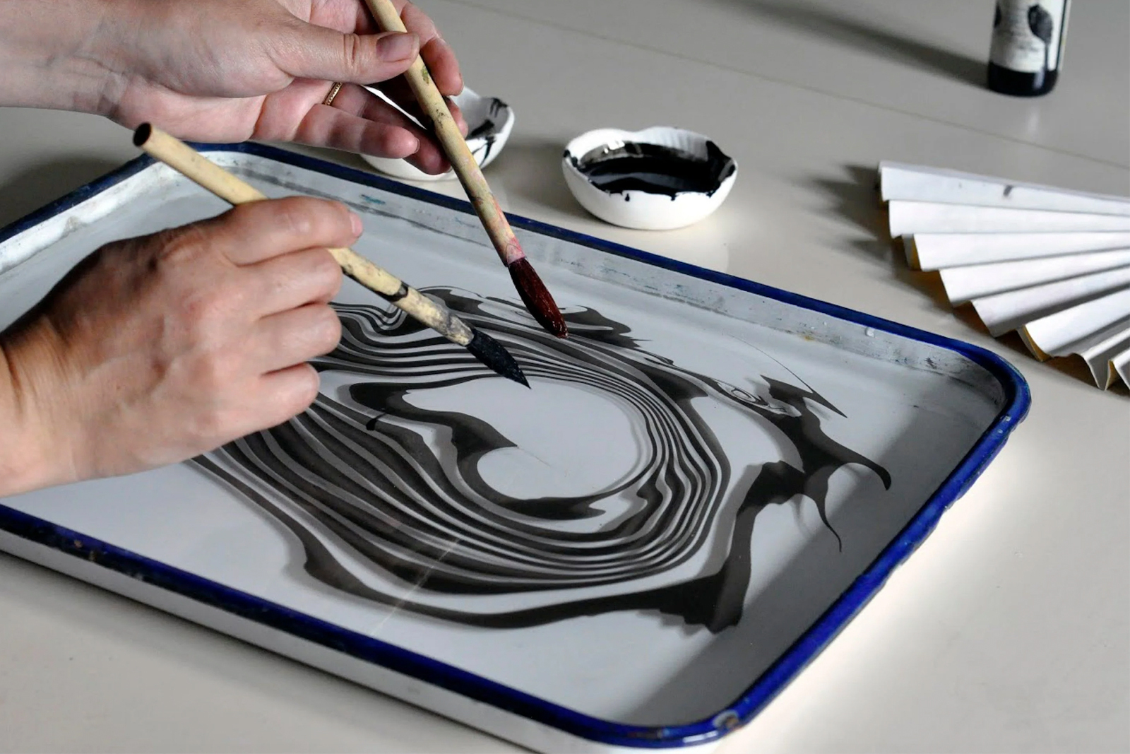 Hands holding brushes over a tray of water with swirls of ink demonstrating paper marbling
