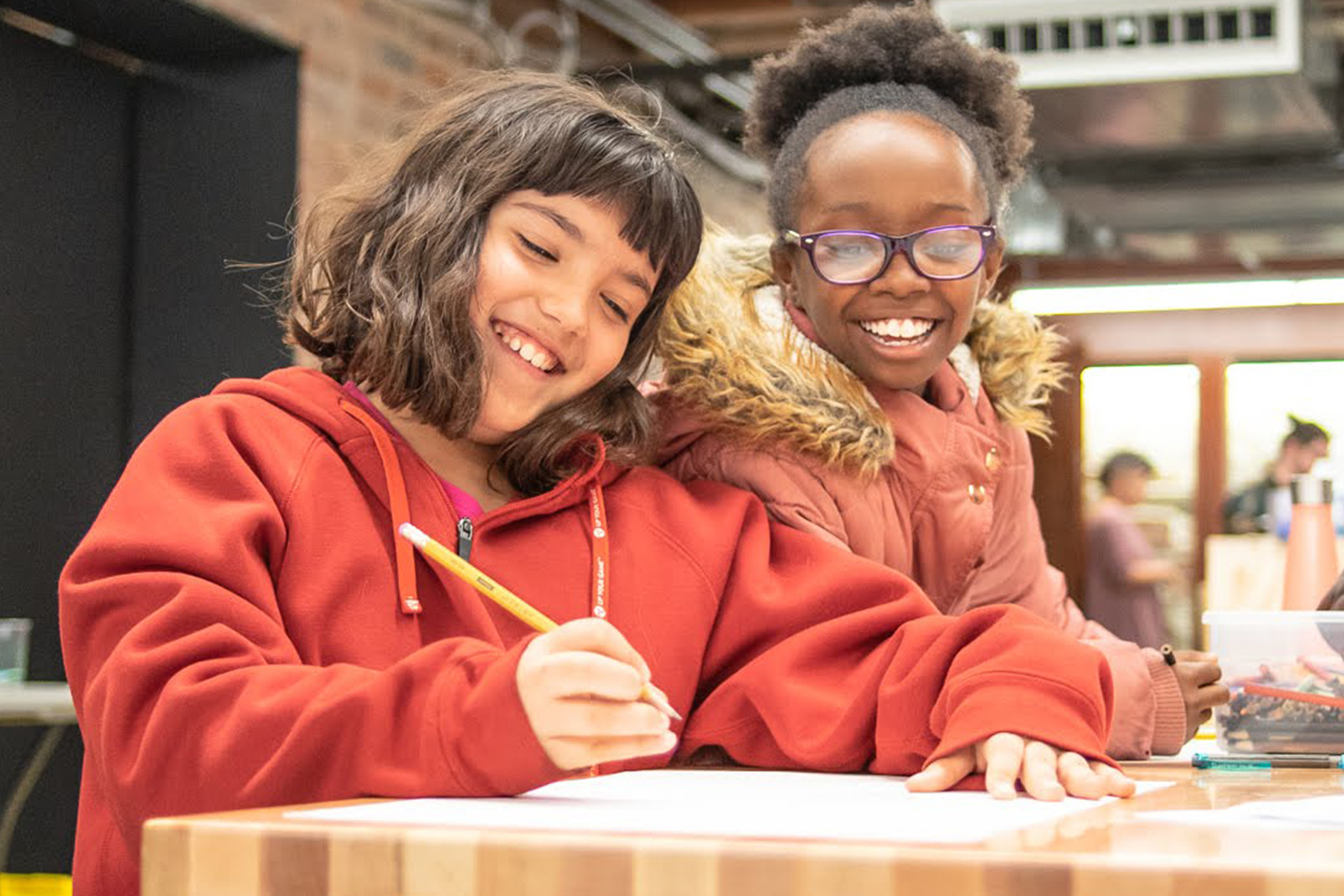 Homepage two young girls smile while working together at a table the girl in the foreground holds a pencil while writing in a notebook
