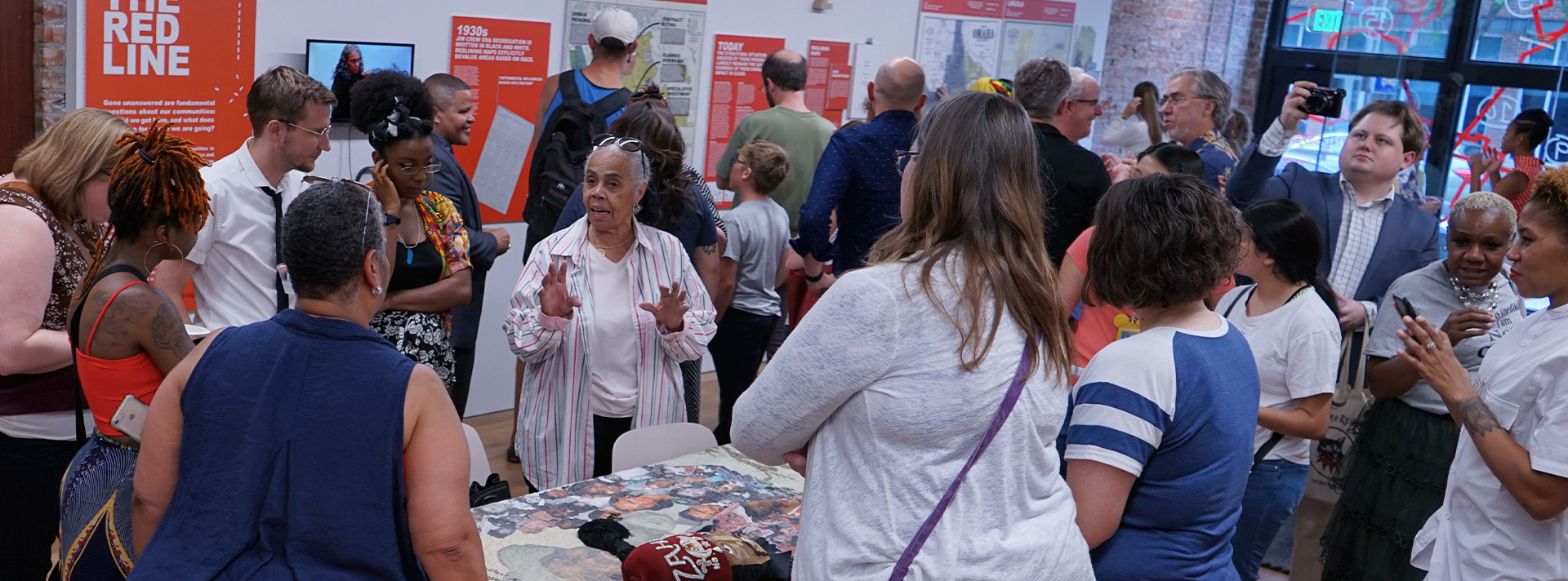 REDLINE BANNER - Dozens of community members gathered in the Undesign the Redline exhibit, several gathered around a community elder telling a story.