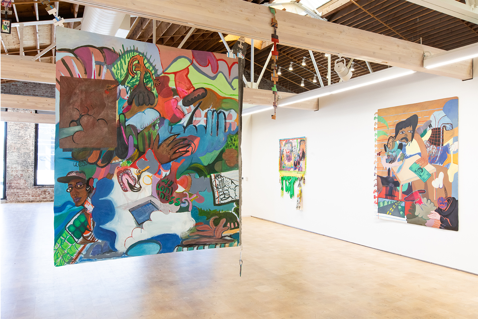 Half gallery view of Rodgers' exhibit with a large unframed canvas handing from the center beam. Canvas is painted is an abstract collage of bright colors, including a small self portrait of the artist peeking out from behind a cloud.