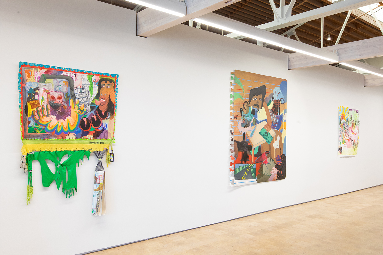 Thalia Rodgers - Three paintings hung on white gallery wall. The work mosts clearly in focus is unframed canvas with strips of green fabric sewn to the bottom. Painting include a face sticking a tongue out and a devilish looking face with red eyes.