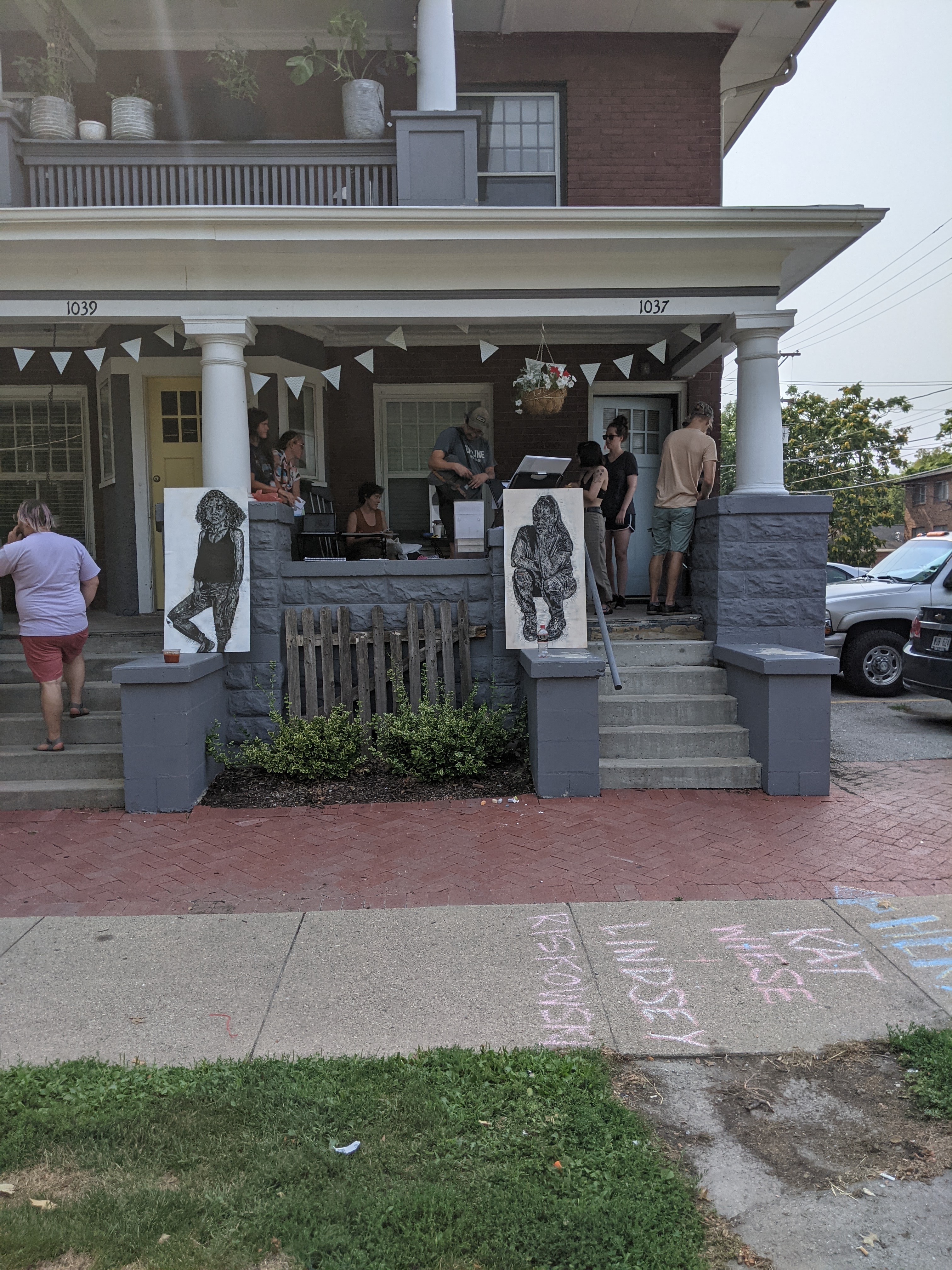 Two Canvases With Illustrations Sit On Two Neighboring Porches While People Gather On The Right Porch