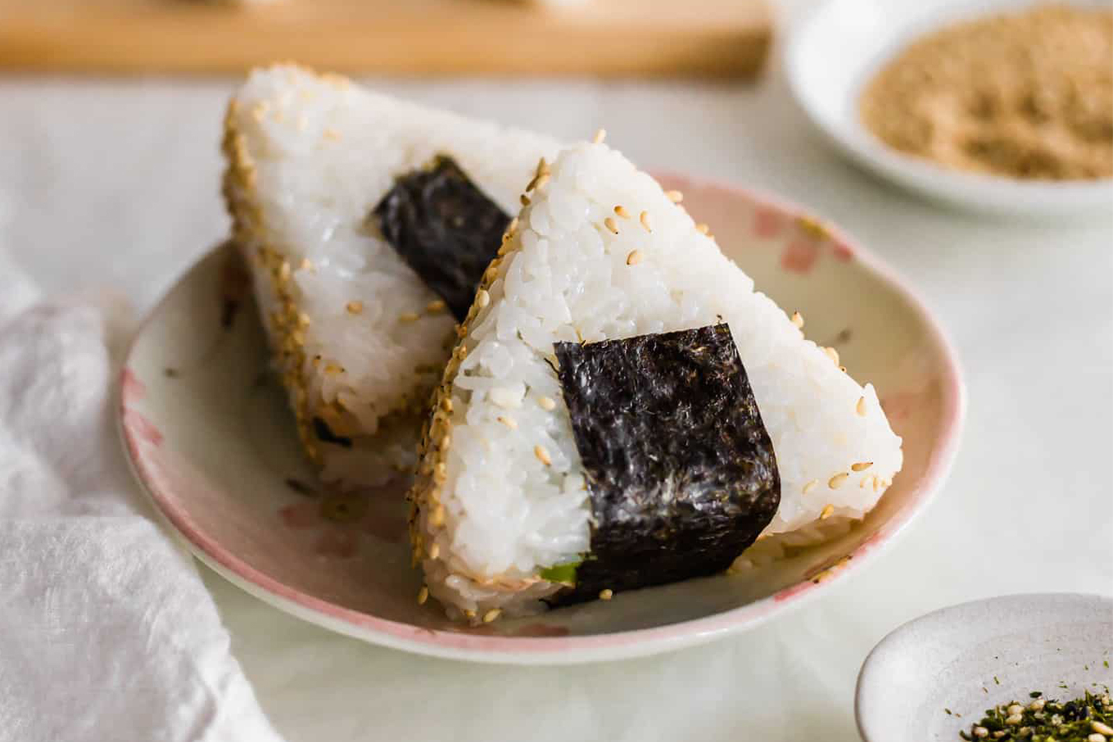 Two onigiri rice balls sit on a small decorative plate with more rice balls out of focus in the background jpg