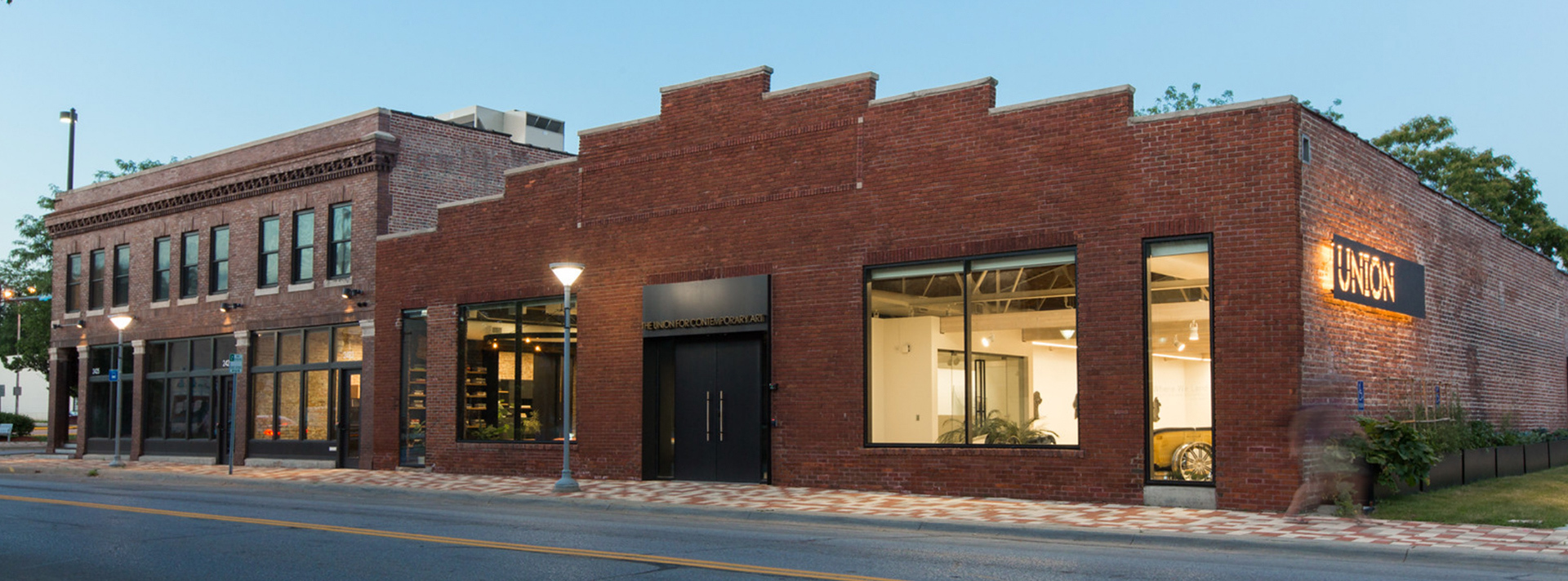 Union Building Banner - Photo of The Union for Contemporary Art taken from North 24th Street. Photo is taken at dusk under a darkening blue sky. The brick building is illuminated from the inside and a black steel Union sign is illuminated on the outside.