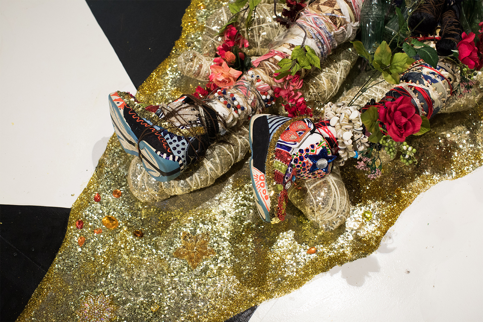 Vanessa German - detail of feet of figure laying on the ground. The feet wear high-top sneakers covered in sequins and paint. Figure is laying on a a triangle of gold glitter against a black and white floor