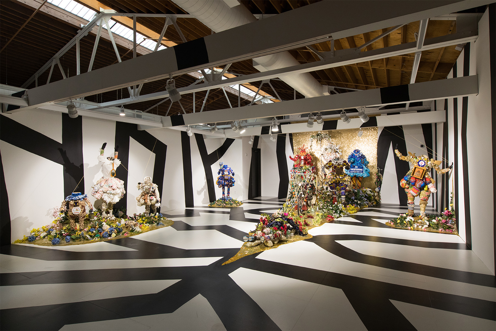 Vanessa German - full view of exhibit. The gallery is painted with black and white stripes filling the walls and floor. 10 life-size human figures are distributed throughout. The feeling is ominous but also whimsical and joyous.