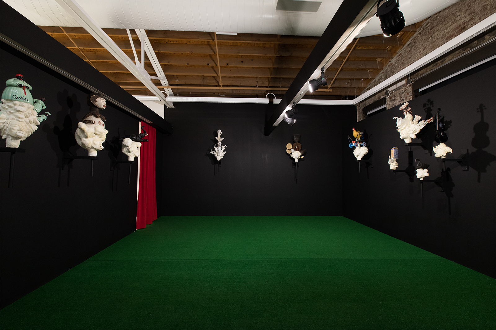 Vanessa German - antechamber to exhibit, black painted walls, green Astroturf on the floor, eight white sculpted heads hanging on the walls, and red curtains leading to the main exhibit.