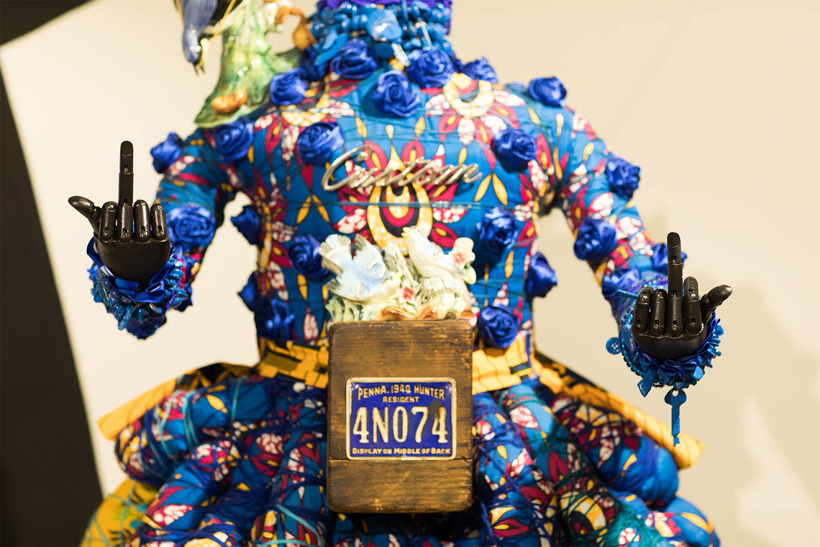 Vanessa German - Detail of figure, wearing a blue assembled dress, a license plate at the navel, and gesturing with two outstretched black hands both showing the middle finger.