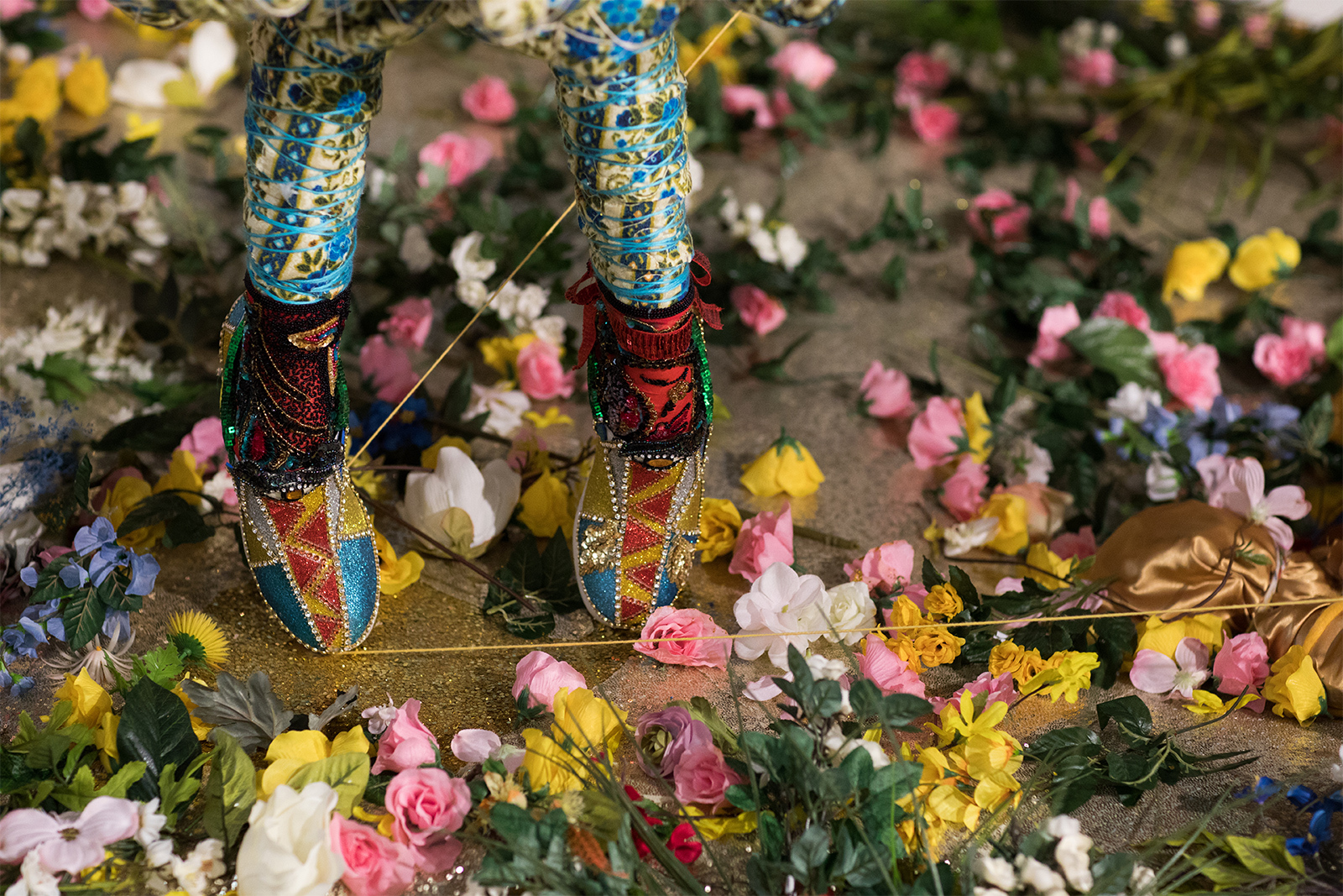 Vanessa German - Detail of a the feet of a figure, standing on a bed of artificial flowers, wearing pointed sequined shoes.