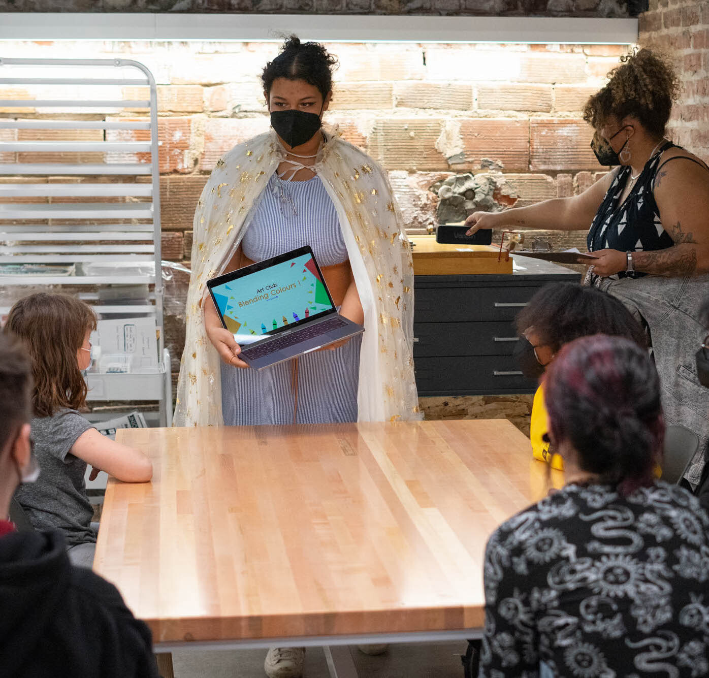 Youth mentor stands at center of a table with young artists looking on