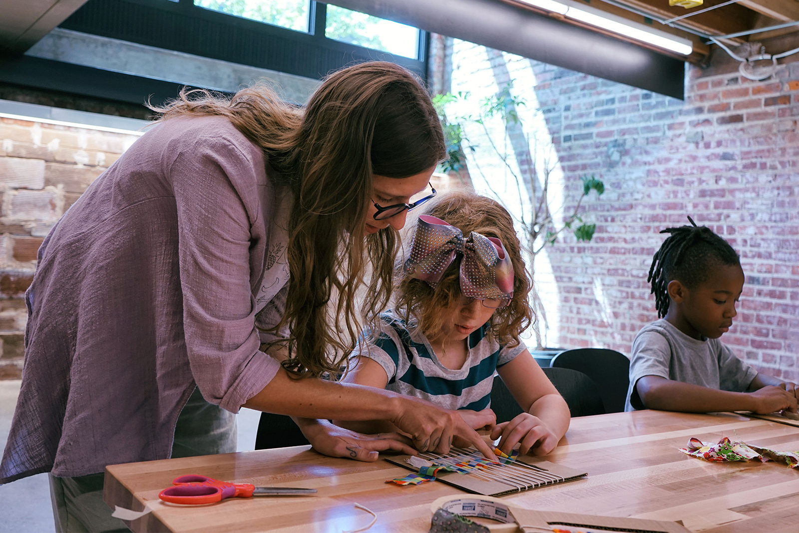 A teaching artist leans over the shoulder of a young girl helping her with a weaving project