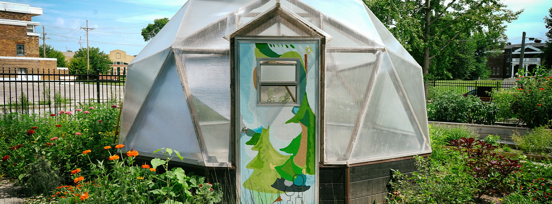 Abundance garden banner - photo of geodesic dome in Union garden. The garden is blooming with flowers and grasses and the door of the dome is painted with a bright, abstract nature scene.