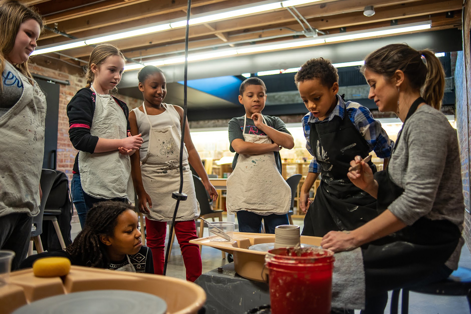 Several young people gather around a pottery wheel where a teaching artsit sits