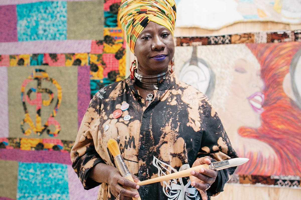 2017 Union Fellow Celeste Butler stands in front of two of her large quilts. She wears a yellow patterned head wrap and artist's smock, holding two paintbrushes. (Union program fellowship-1)