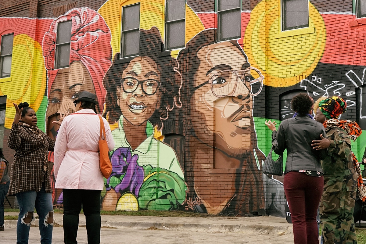 Union Program Neighborhood Arts - Community members in conversation stand in front of the mural "Ancestor, Identity, and the Seed" featuring portraits of 3 Black people of different ages against the Pan-African flag.