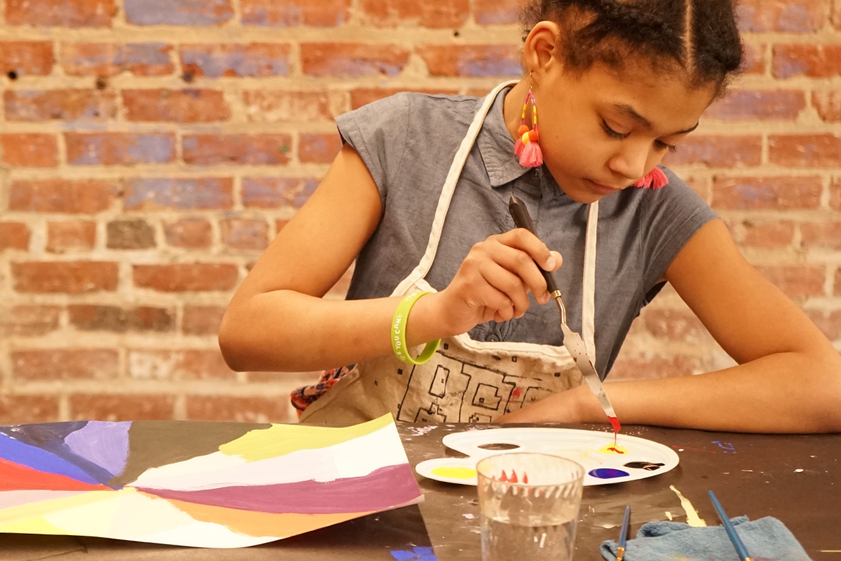 Union Program Youthengagement - Young artist at work, seated in front of a brick wall wearing an apron and mixing paints in a palette.