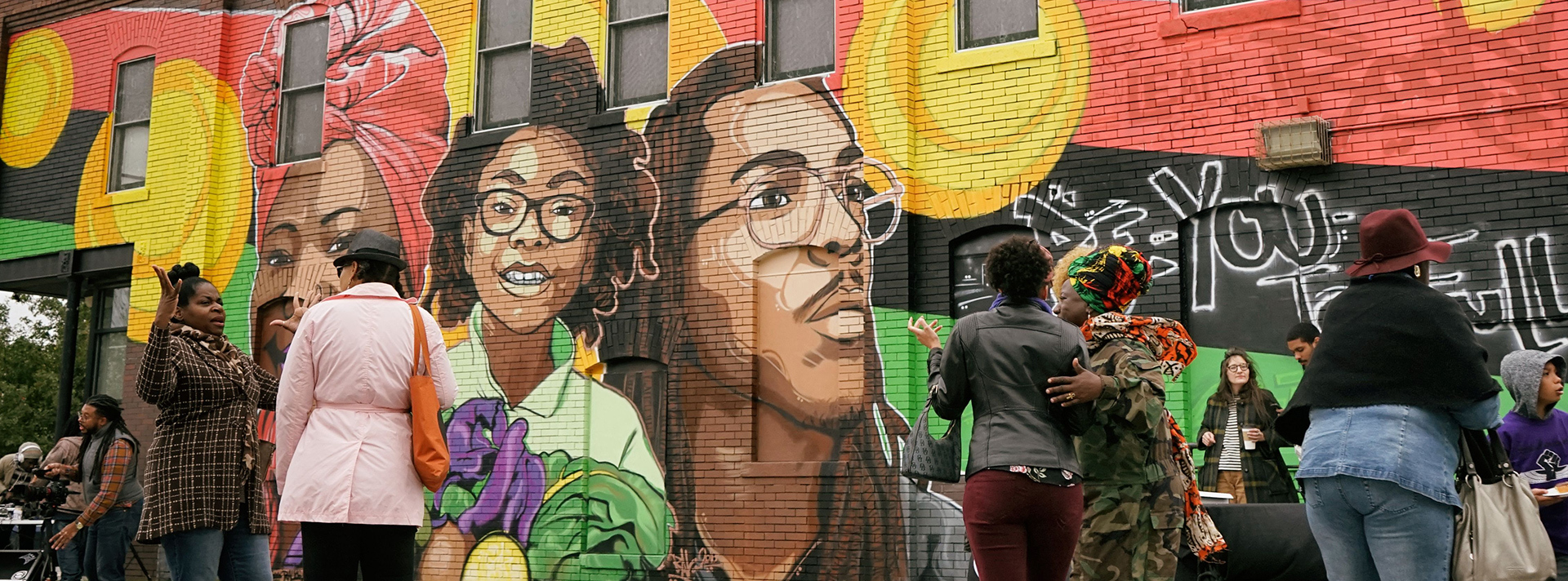 Header-Neighborhood Arts - Community members in conversation stand in front of the mural "Ancestor, Identity, and the Seed" featuring portraits of 3 Black people of different ages against the Pan-African flag.