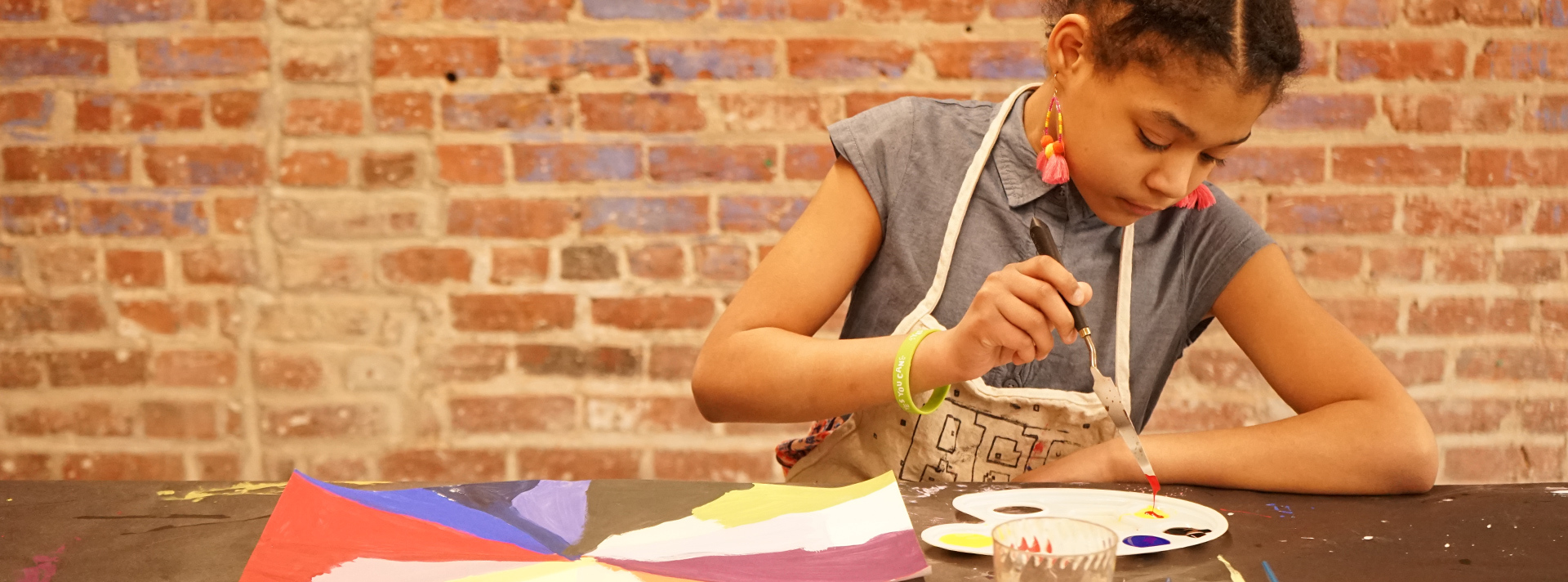 Youth engagement Header - Young artist at work, seated in front of a brick wall wearing an apron and mixing paints in a palette.